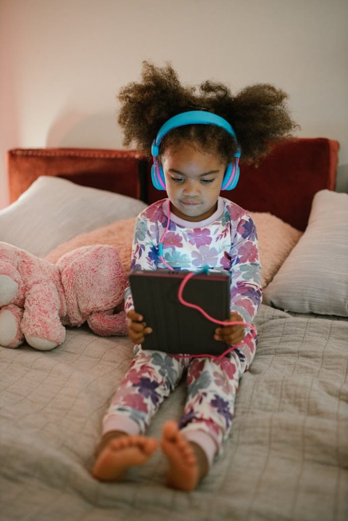 Young girl with headphones using a tablet on bed, surrounded by pillows and stuffed toys.